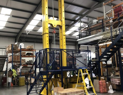yellow Vertical drop test machine - approx 3 stories tall. black stair case leads up to the platform half way up for the operator to load samples on.